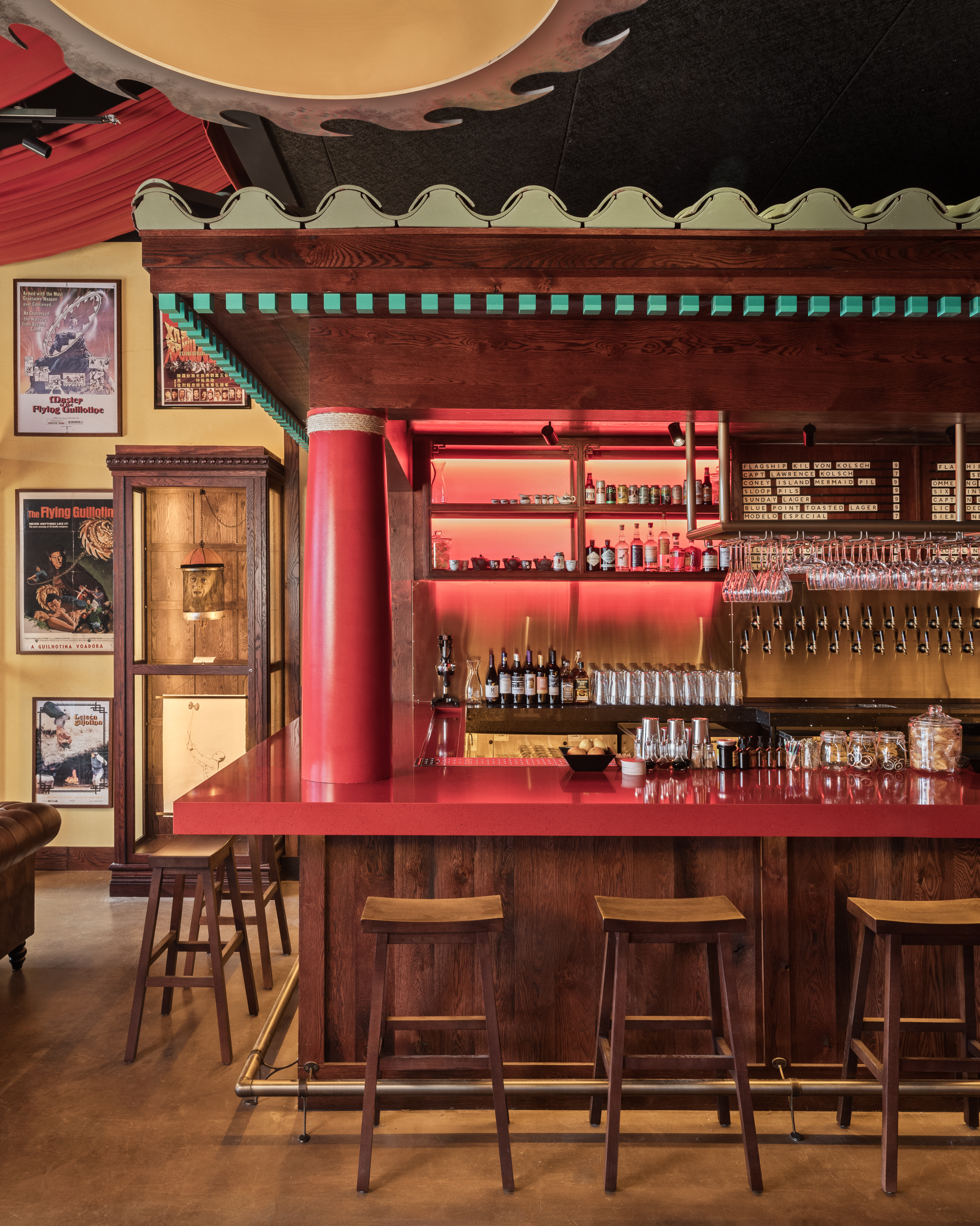 Front shot of a bar corner with red columns and countertops, wooden paneling, and a wooden top section with green trim and light green roofing. Hanging above the bar is the edge of a flying guillotine, as seen in the movie Master of The Flying Guillotine, hung from the ceiling. The bar corner is surrounded by wooden stools with square seats.  Behind the bar counter are clean glasses hung upside-down and several bottles of spirits/liquors. The walls are a yellow off-white color that have wooden paneling and a chair rail. On the wall to the right of the bar are a few posters for Master of The Flying Guillotine and a (hopefully fake) decapitated head hanging from a chain inside a wooden case.