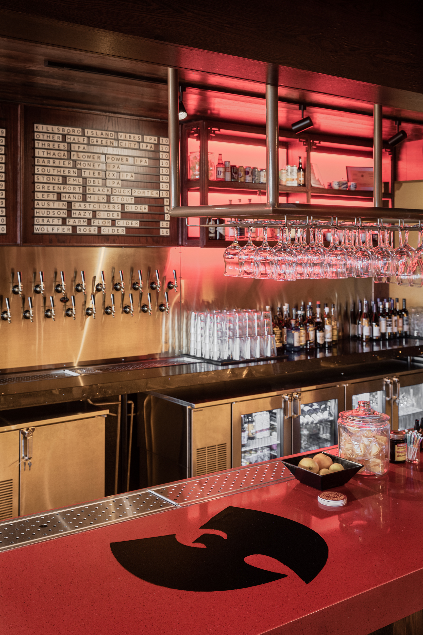 A shot of a red bar's surface with a black Wu-Tang Clan "W" logo in the center of the bar's surface. Sitting on the bar surface are a bowl of blood oranges, a jar of fortune cookies, and other assorted bar goodies to the right. Behind the bar are several beer taps, a menu of several drinks, and neatly organized sets of clean glasses stacked upside down, and various bottles of liquor/spirits.