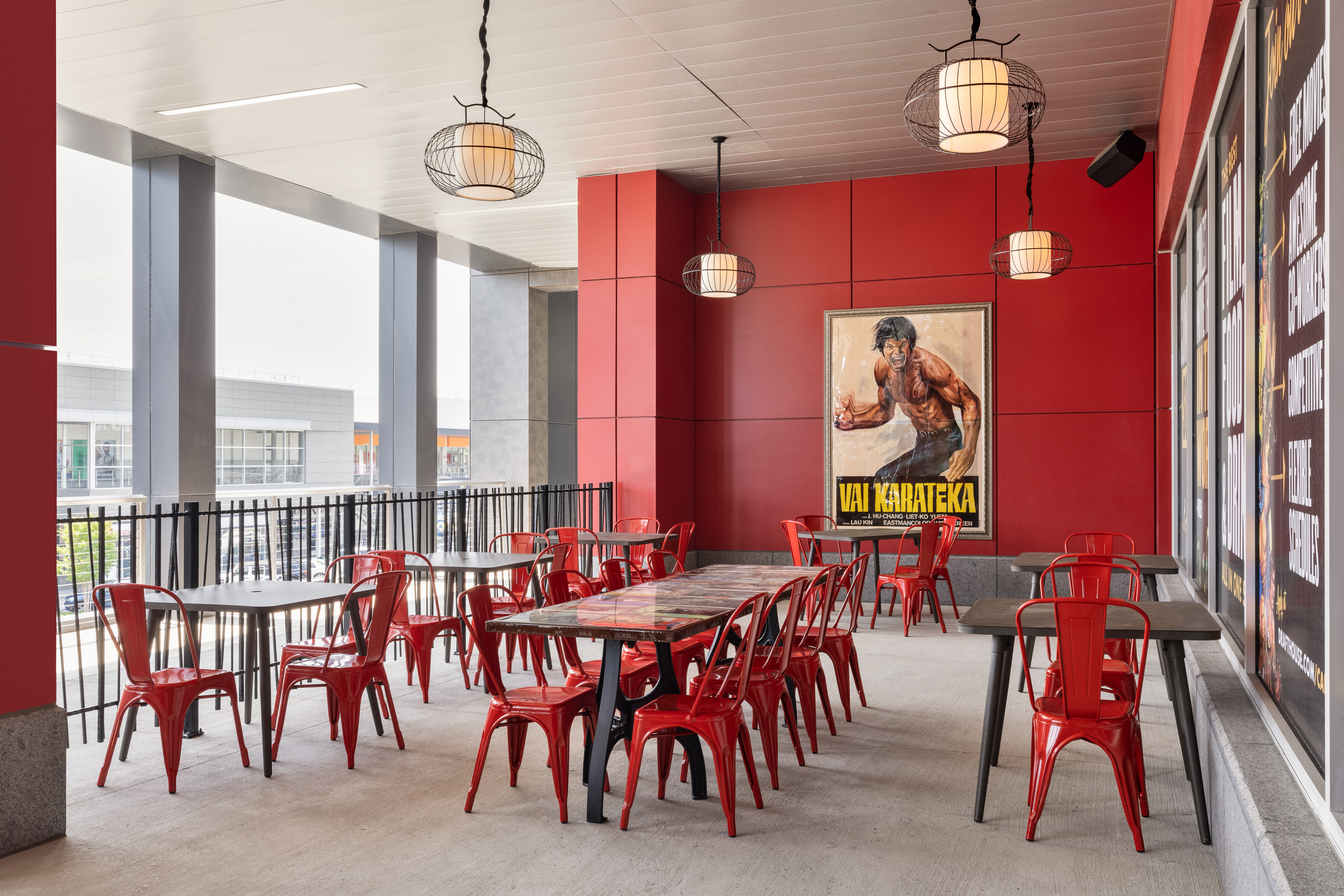 An outdoor patio with several tables with seating. On a red wall behind the tables is a framed movie poster for "Val Karateka"
