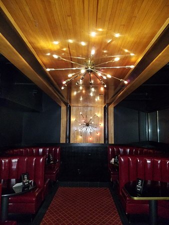 A ceiling that looks like it was made of wood from a bowling lane adorns a chandelier below a four dark red colored vinyl dining seats next to gold colored tables, surrounded by dark teal walls.