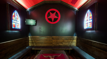 An occult temple themed room with a crimson red ceiling, black walls surrounded by stainglass windows of an inverted cross, shaped like an upside down shield. On the wall, just above in the center is a solid red pentagram with the design of a goat within the five-pointed star. In all three sides of the shown room are wooden pews.