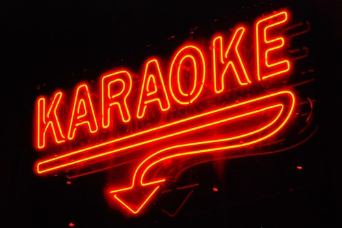 A bright orange neon sign of the word "KARAOKE", with an underline that become a trailing line that becomes an arrow pointing down, is glowing.