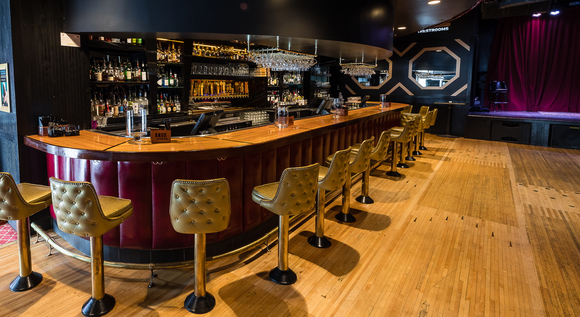 A bar with a glossed wooden countertop and a bar wall with red vinyl cushioning, surrounded by cushion gold bar seats. Behind the bar are various bottles of liquors, spirits and mixers. Behind the bar's seating, to the right, is a wooden dancefloor with bowling lane markers in some spots.