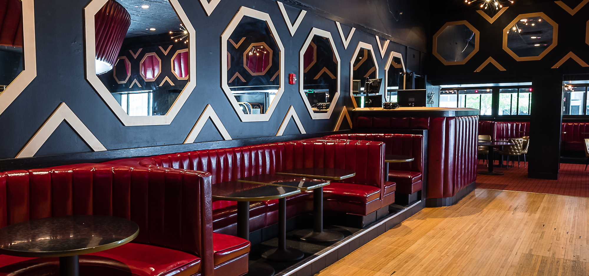 Several dining booths with dark gold colored tables and dark red vinyl seats. On the black walls are patterns of hexagon mirrors and vertical triangles, both with white trim.