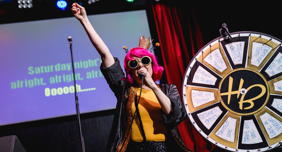 A girl wearing a bright short-haired pink wig with curly antennas that appears to be made of pipe cleaners and a crown on top of her head, round dark sunglasses, orange shirt and short-sleeved black overcoat raises one fist in the air while hold a microphone in the other is singing on stage. To the right of her, directly behind, is a spinner wheel with several songs listed such as "Danger Zone", "Eye of The Tiger", and "Hungry Like The Wolf". The spinner's pointer is on "Singer's Choice". On the stage's screen are the song lyrics "Saturday night's alright, alright, alright, Oooooh......"