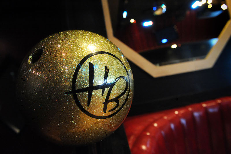 A fixed glittering gold bowling ball with a stylized "HB" logo in black emblazoned on it. In the background is a red vinyl booth below a hexagon-shaped mirror with white trim.